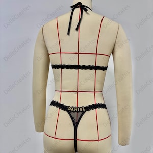 Puede incluir: Conjunto de lencer&iacute;a negra con ribete de encaje y el nombre "DANIEL" en letras doradas. El conjunto incluye un top halter, una banda en la cintura y un tanga. La lencer&iacute;a se exhibe en un maniqu&iacute;.