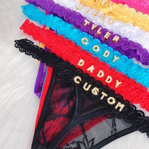 May include: A stack of lace thong underwear in black, red, blue, purple, white, and yellow. Each pair features a scalloped lace waistband adorned with gold-toned rhinestone letters spelling out custom names and words like DADDY and CUSTOM.