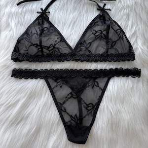 Puede incluir: Conjunto de lencer&iacute;a negra con un sujetador triangular con tirantes anudados y un tanga a juego. El conjunto est&aacute; hecho de encaje transparente con estampado floral y bordes festoneados. El sujetador tiene peque&ntilde;os lazos en los v&eacute;rtices.