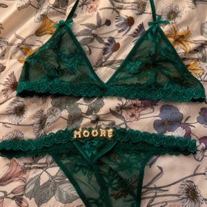 May include: A dark green lingerie set consisting of a triangle bralette and matching thong. Both pieces feature sheer floral lace, scalloped heart-patterned trim, and small bows. The thong displays the name MOORE in sparkling rhinestone letters.