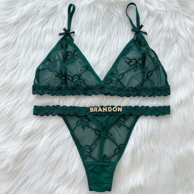 Puede incluir: Conjunto de lencer&iacute;a verde esmeralda con un sujetador triangular y tanga a juego. El conjunto es de encaje transparente con motivos florales y delicados adornos. El tanga tiene el nombre "BRANDON" en letras doradas. El sujetador tiene peque&ntilde;os lazos negros.