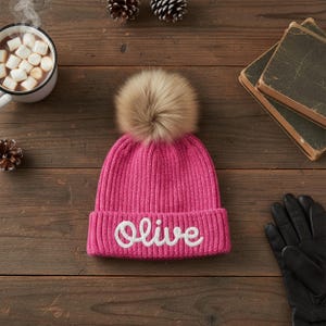 Puede incluir: Un gorro de punto rosa brillante con un gran pompón marrón claro y esponjoso. La palabra "Olive" está bordada en blanco en la parte delantera. El gorro está sobre una superficie de madera, con una taza de chocolate caliente y guantes negros.