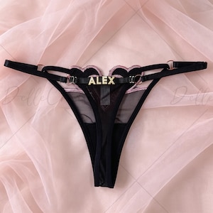 May include: Black thong underwear with a sheer mesh front panel. The waistband features a heart-shaped design with pink trim and the name "ALEX" in gold lettering. The straps are black with gold-colored rings.