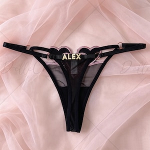 May include: Black thong underwear featuring a sheer mesh panel, heart-shaped embroidery with pink trim, and thin adjustable straps with gold-tone metal rings. A central horizontal strap displays the name ALEX in gold-tone block lettering.