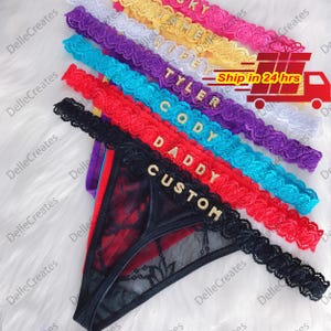 May include: A stack of black, red, blue, purple, white, yellow, and pink lace thongs with gold glitter lettering. The lettering reads "Custom", "Daddy", "Cody", "Tyler", "Jamie", and "Sky".