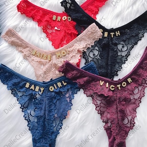 May include: Five pairs of lacy underwear with gold lettering. The lettering on the underwear reads "BROD", "BAISE MOI", "ETH", "BABY GIRL", and "VICTOR".