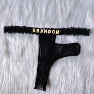 May include: A black lace thong featuring a scalloped elastic waistband adorned with the name BRANDON in gold-toned, rhinestone-encrusted letters. The garment is crafted from a sheer, floral-patterned black lace fabric with a solid black gusset.