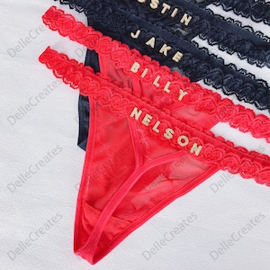 May include: Close-up of three thong underwear pieces in red and black lace. The red thong has the name "NELSON" spelled out in gold letters. The black thong has the name "JAKE" spelled out in gold letters. The underwear has a delicate lace trim.
