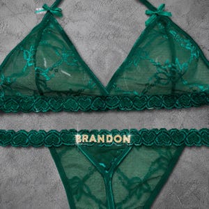 Puede incluir: Un conjunto de lencería de encaje verde oscuro compuesto por un bralette triangular y una tanga a juego. El conjunto presenta un patrón de encaje floral, bordes en forma de corazón y pequeños lazos. La palabra BRANDON aparece en letras doradas.