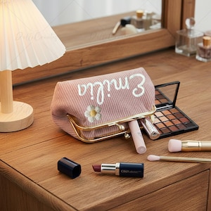 Puede incluir: Un neceser de maquillaje de pana rosa con el nombre "Emilia" bordado en escritura blanca y una margarita. La bolsa está abierta, revelando barras de labios y una paleta de maquillaje. Un labial y brochas de maquillaje están sobre la superficie de madera.