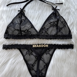 Puede incluir: Conjunto de lencer&iacute;a de encaje negro con un sujetador triangular con tirantes halter y un tanga a juego. El conjunto tiene patrones florales y la palabra "BRANDON" en letras doradas en el tanga. Se muestra sobre una superficie blanca y esponjosa.