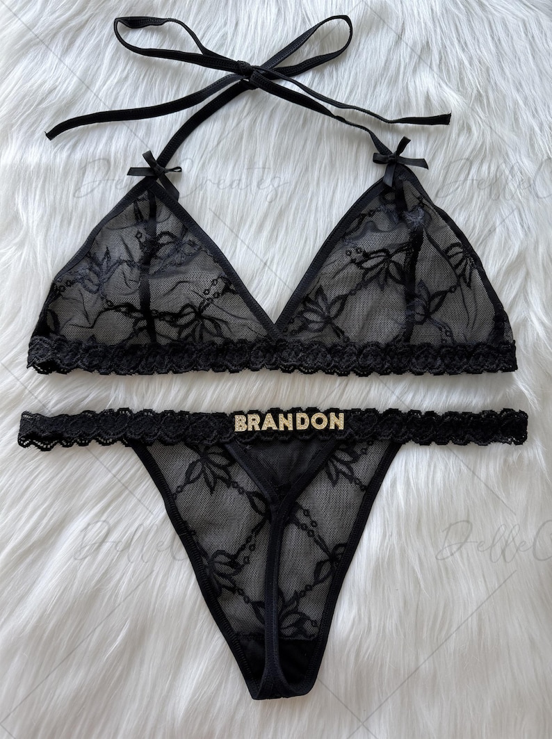 Puede incluir: Conjunto de lencer&iacute;a de encaje negro con un sujetador triangular con tirantes halter y un tanga a juego. El conjunto tiene patrones florales y la palabra "BRANDON" en letras doradas en el tanga. Se muestra sobre una superficie blanca y esponjosa.