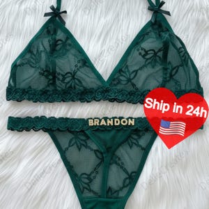 May include: Emerald green lingerie set with floral lace detailing. The set includes a triangle bralette with bow accents and a matching thong. The word "BRANDON" is spelled out in gold letters on the waistband.