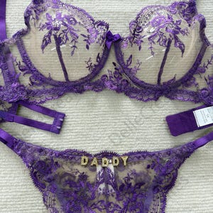May include: A purple lingerie set featuring a bra and thong. The bra has floral embroidery and adjustable straps. The thong has the word "DADDY" spelled out in gold letters. The set is made of sheer fabric.