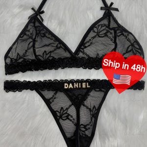 Puede incluir: Conjunto de lencer&iacute;a de encaje negro con un sujetador triangular con peque&ntilde;os lazos y un tanga a juego. El tanga tiene el nombre "DANIEL" en letras doradas. El conjunto est&aacute; hecho de una tela transparente con estampado floral.