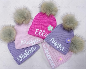 May include: Five colorful knit beanies with faux fur pom-poms. The beanies are in shades of pink and purple, each with a name embroidered in white script. Each hat has a small flower accent. The names on the hats are Elle, Maya, Vivian, Nova, and Cameron.