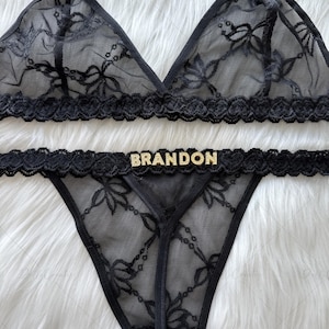 Puede incluir: Conjunto de lencer&iacute;a de encaje negro. El sujetador triangular tiene detalles de lazo, y la tanga a juego lleva el nombre "BRANDON" en letras doradas. El conjunto est&aacute; hecho de encaje transparente con estampado floral.