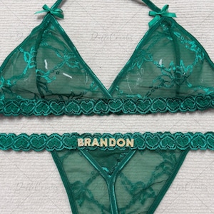 May include: A two-piece emerald green lingerie set featuring a sheer mesh triangle bralette with floral embroidery, small bows at the straps, and a matching thong with a heart-patterned lace waistband displaying the name BRANDON in gold.