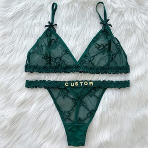 Puede incluir: Un conjunto de lencería verde oscuro que incluye un bralette y una tanga. El bralette tiene tirantes finos con lazos pequeños y un patrón de encaje floral. La tanga presenta una cintura de encaje festoneado con la palabra CUSTOM en letras doradas.