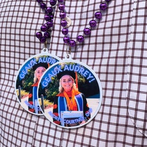 May include: Two white graduation medallions with a photo of a smiling graduate in a cap and gown. The medallions have the text "GEAUX AUDREY!" in blue and the graduate's name. Purple beaded necklace hangs above the medallions.