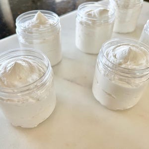 May include: Several clear plastic jars filled with white, whipped cream-like substance. The jars are arranged on a white marble surface. The contents of the jars have a fluffy, textured appearance, suggesting a cosmetic or food product.