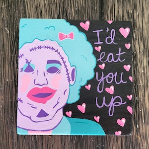 May include: Square wooden art piece featuring a stylized illustration of a character with blue hair, pink lips, and stitched facial details. The text "I'd eat you up" is written in purple with pink hearts on a black background.