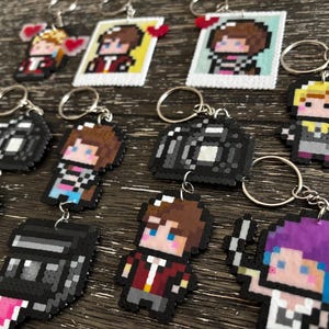 May include: A collection of pixel art keychains featuring various characters and cameras. The designs include figures with brown, blonde, and purple hair, alongside black camera shapes and square frames with red heart accents, all attached to metal rings.