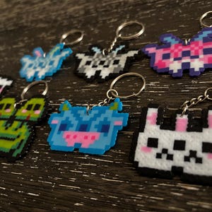 May include: A collection of pixel art keychains in various colors and designs. The keychains feature animal and creature shapes, including a frog, a cow, and a cat. Each keychain has a silver keyring attached.