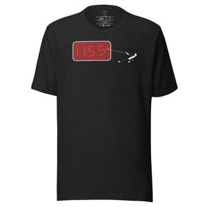 Puede incluir: Camiseta gris oscuro con un reloj digital rojo que muestra "11:55" y un diseño minimalista de un muñeco de palo. Una línea conecta el reloj con la figura, sugiriendo un concepto de despertador.