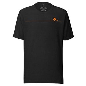 May include: Dark gray t-shirt with an orange hockey player graphic. The design features a horizontal line and a hockey player in action. The shirt has a crew neck and short sleeves. The shirt has a tag at the back of the neck.