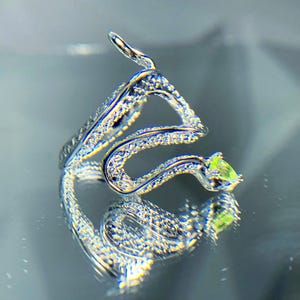 May include: A silver snake ring with a winding design, set with small, clear gemstones. The ring has a pear-shaped, light green gemstone at the head. The ring is displayed on a reflective surface.