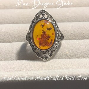 K&ouml;nnte beinhalten: Ein silberner Ring mit einem ovalen Bernstein, der in einem filigranen Design gefasst ist. Der Bernstein hat eine warme, honigartige Farbe und enth&auml;lt Einschl&uuml;sse. Der Ring ist handgefertigt und tr&auml;gt den Text "Muse Designer Studio" und "Real Gemstone, HandCrafted With Love".