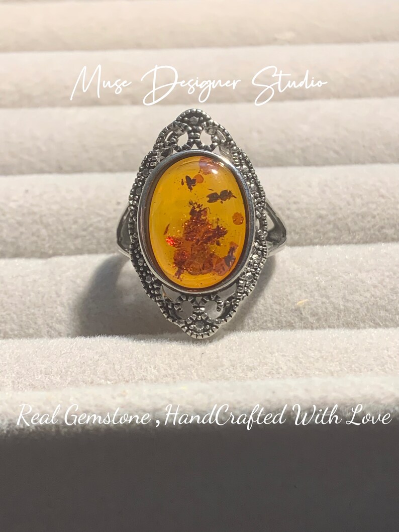 K&ouml;nnte beinhalten: Ein silberner Ring mit einem ovalen Bernstein, der in einem filigranen Design gefasst ist. Der Bernstein hat eine warme, honigartige Farbe und enth&auml;lt Einschl&uuml;sse. Der Ring ist handgefertigt und tr&auml;gt den Text "Muse Designer Studio" und "Real Gemstone, HandCrafted With Love".