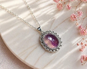 May include: Silver-toned necklace with an oval pendant featuring a purple and pink gemstone. The pendant is framed by a decorative, scalloped border. The necklace rests on a white, marble-like surface, with pink flowers in the background.