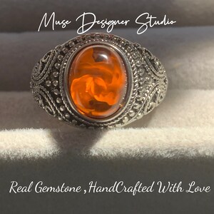 K&ouml;nnte beinhalten: Ein silberner Ring mit einem orangefarbenen Edelstein, der in einem dekorativen Filigranmuster gefasst ist. Der Ring wird in einer wei&szlig;en Schachtel mit dem Text "Muse Designer Studio" und "Real Gemstone, HandCrafted With Love" pr&auml;sentiert.