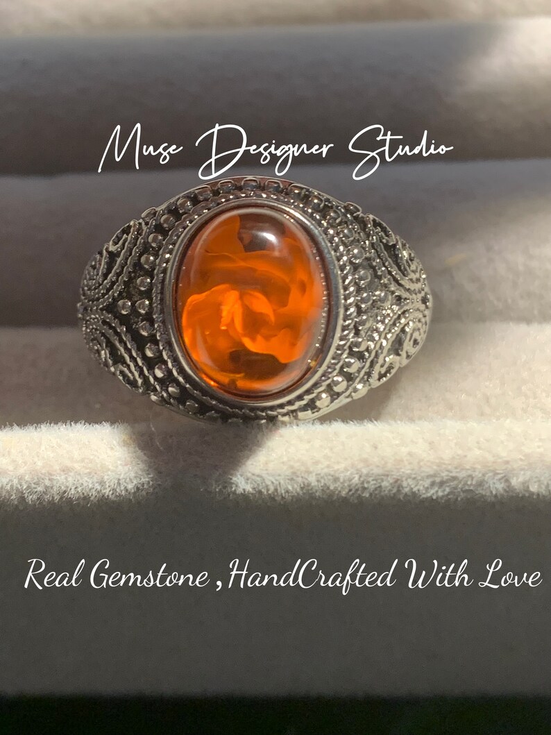 K&ouml;nnte beinhalten: Ein silberner Ring mit einem orangefarbenen Edelstein, der in einem dekorativen Filigranmuster gefasst ist. Der Ring wird in einer wei&szlig;en Schachtel mit dem Text "Muse Designer Studio" und "Real Gemstone, HandCrafted With Love" pr&auml;sentiert.