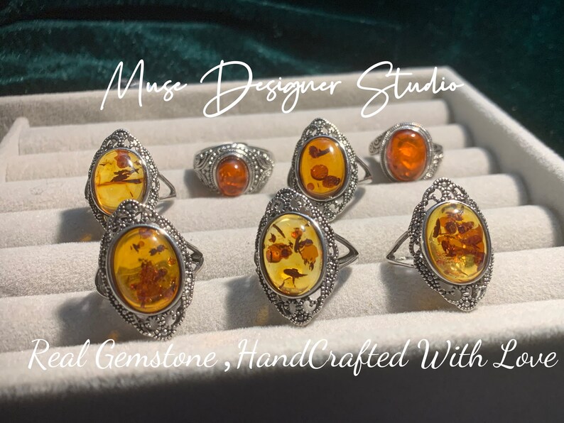 K&ouml;nnte beinhalten: Sechs Silberringe mit ovalen Bernstein-Edelsteinen. Die Ringe haben komplizierte Filigranarbeiten. Der Text "Muse Designer Studio" und "Real Gemstone, HandCrafted With Love" ist im Bild sichtbar.