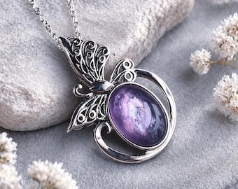 May include: Silver-toned butterfly pendant necklace with a purple oval-shaped stone. The butterfly design features intricate filigree wings and a delicate chain. The necklace is displayed on a gray surface with white flowers.