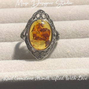 K&ouml;nnte beinhalten: Ein silberner Ring mit einem ovalen Bernstein. Der Ring hat ein filigranes Design und der Bernstein enth&auml;lt Einschl&uuml;sse. Der Ring befindet sich in einer wei&szlig;en Schachtel mit dem Text "Muse Designer Studio" und "Real Gemstone, HandCrafted With Love".