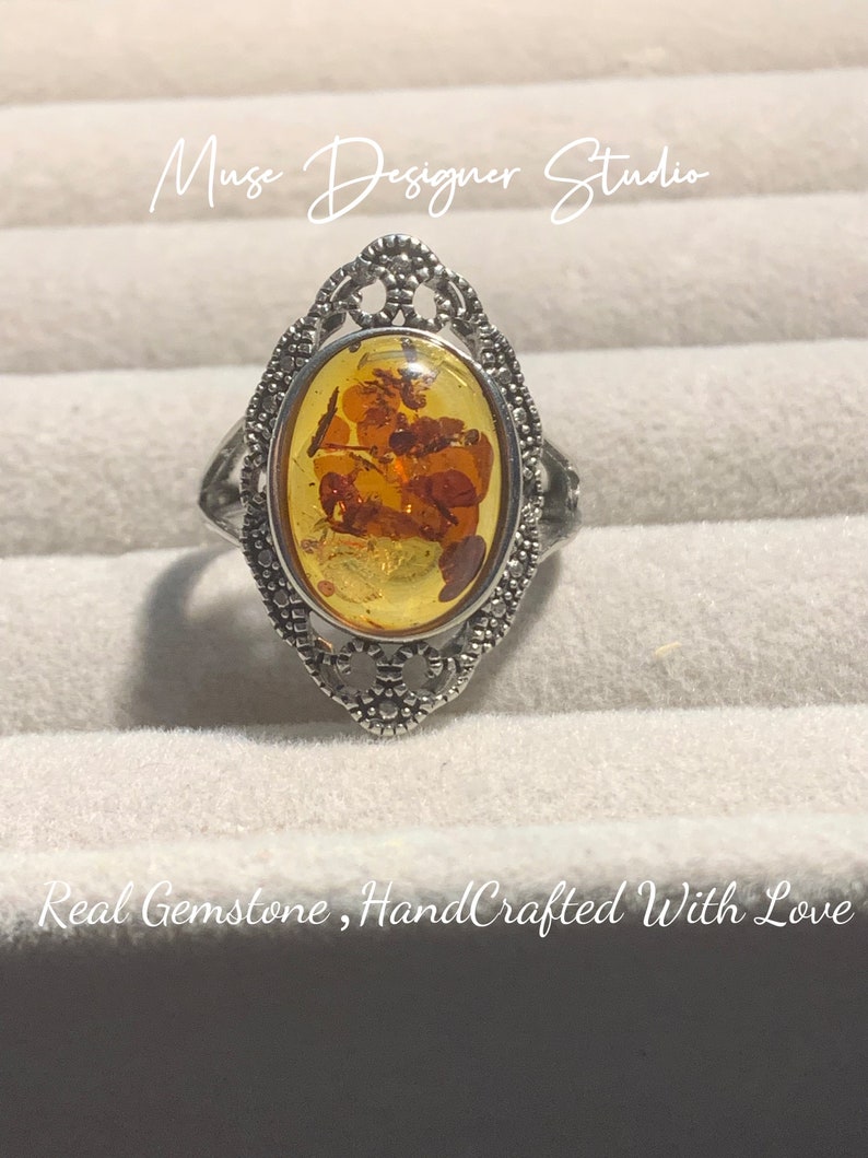 K&ouml;nnte beinhalten: Ein silberner Ring mit einem ovalen Bernstein. Der Ring hat ein filigranes Design und der Bernstein enth&auml;lt Einschl&uuml;sse. Der Ring befindet sich in einer wei&szlig;en Schachtel mit dem Text "Muse Designer Studio" und "Real Gemstone, HandCrafted With Love".