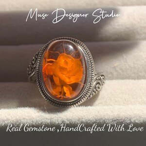 K&ouml;nnte beinhalten: Ein silberner Ring mit einem ovalen, orangefarbenen Edelstein, der in einer silbernen Fassung sitzt. Der Edelstein zeigt ein detailliertes Rosenmotiv. Der Ring hat ein dekoratives Schriftrollenmuster am Band.