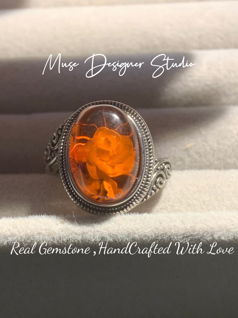 K&ouml;nnte beinhalten: Ein silberner Ring mit einem ovalen, orangefarbenen Edelstein, der in einer silbernen Fassung sitzt. Der Edelstein zeigt ein detailliertes Rosenmotiv. Der Ring hat ein dekoratives Schriftrollenmuster am Band.