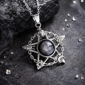 May include: Silver pendant necklace with a pentagram design and a central dark grey stone. The pendant features chain links, skulls, and crescent moon details. The necklace is displayed on a dark, textured surface.