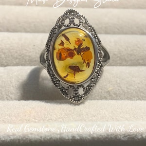 K&ouml;nnte beinhalten: Ein silberner Ring mit einem ovalen Bernstein, der in einem filigranen Design gefasst ist. Der Bernstein hat ein florales Muster mit Einschl&uuml;ssen. Der Ring befindet sich auf einem wei&szlig;en Hintergrund.