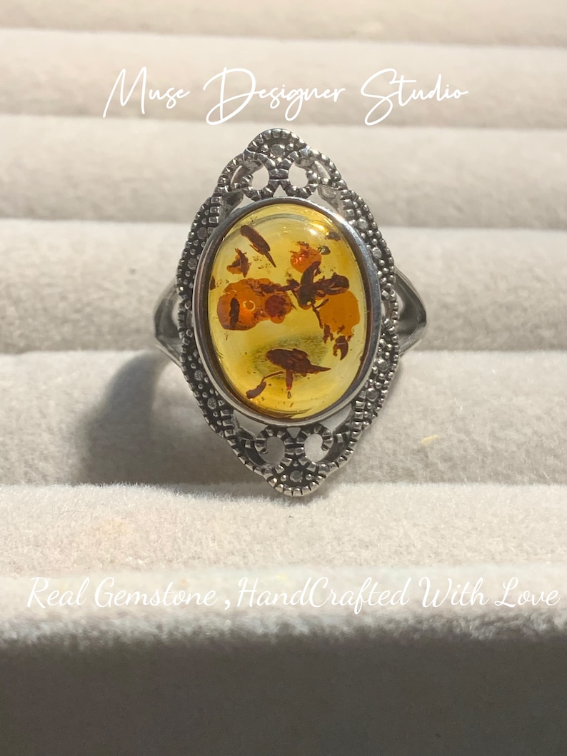 K&ouml;nnte beinhalten: Ein silberner Ring mit einem ovalen Bernstein, der in einem filigranen Design gefasst ist. Der Bernstein hat ein florales Muster mit Einschl&uuml;ssen. Der Ring befindet sich auf einem wei&szlig;en Hintergrund.