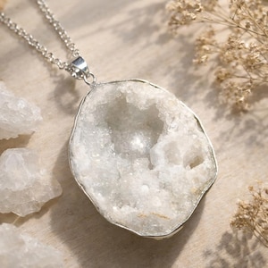 May include: A silver-toned necklace featuring a large, irregular-shaped pendant. The pendant has a silver border and is filled with a sparkling, white crystal formation. The background includes small, white crystal fragments and dried flowers.