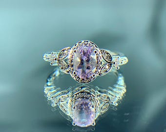 May include: A silver ring with a large oval-shaped purple gemstone surrounded by smaller clear gemstones. The ring has a delicate floral design.