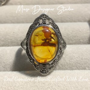 K&ouml;nnte beinhalten: Ein silberner Ring mit einem ovalen Bernstein, der in einem filigranen Design gefasst ist. Der Ring wird in einer wei&szlig;en Schachtel mit dem Text "Muse Designer Studio" und "Real Gemstone, HandCrafted With Love" pr&auml;sentiert.