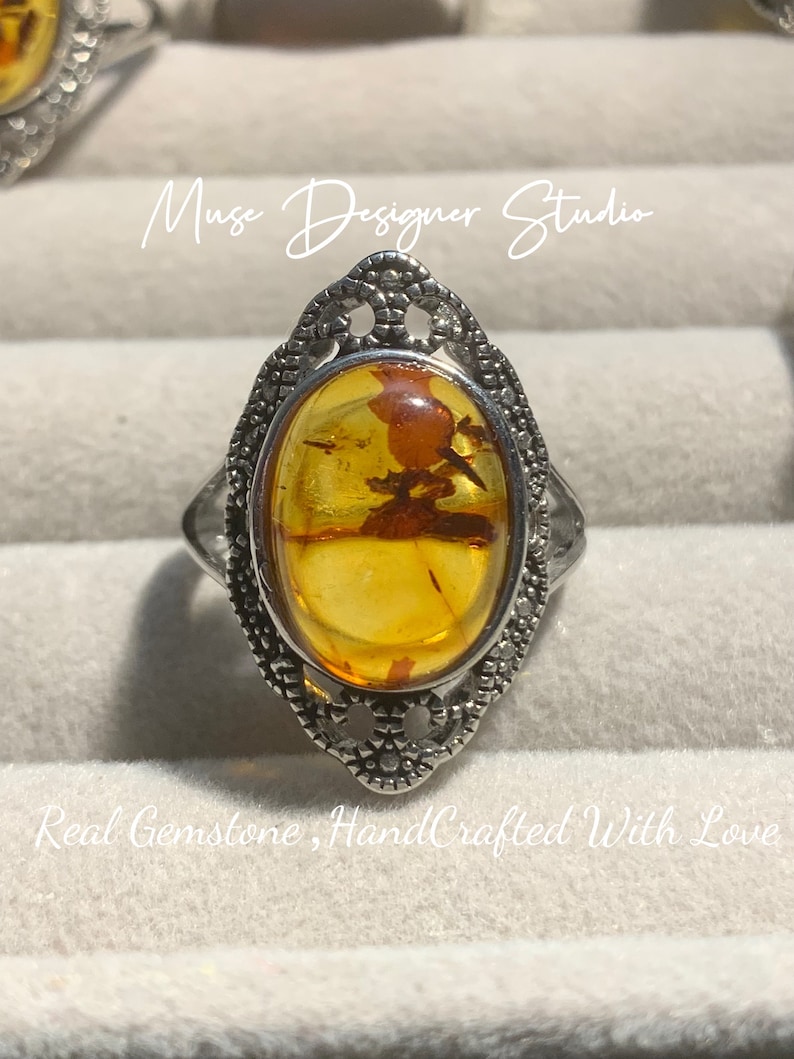 K&ouml;nnte beinhalten: Ein silberner Ring mit einem ovalen Bernstein, der in einem filigranen Design gefasst ist. Der Ring wird in einer wei&szlig;en Schachtel mit dem Text "Muse Designer Studio" und "Real Gemstone, HandCrafted With Love" pr&auml;sentiert.