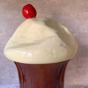 May include: A ceramic ice cream sundae cookie jar with a brown base and a white top with a red cherry on top.